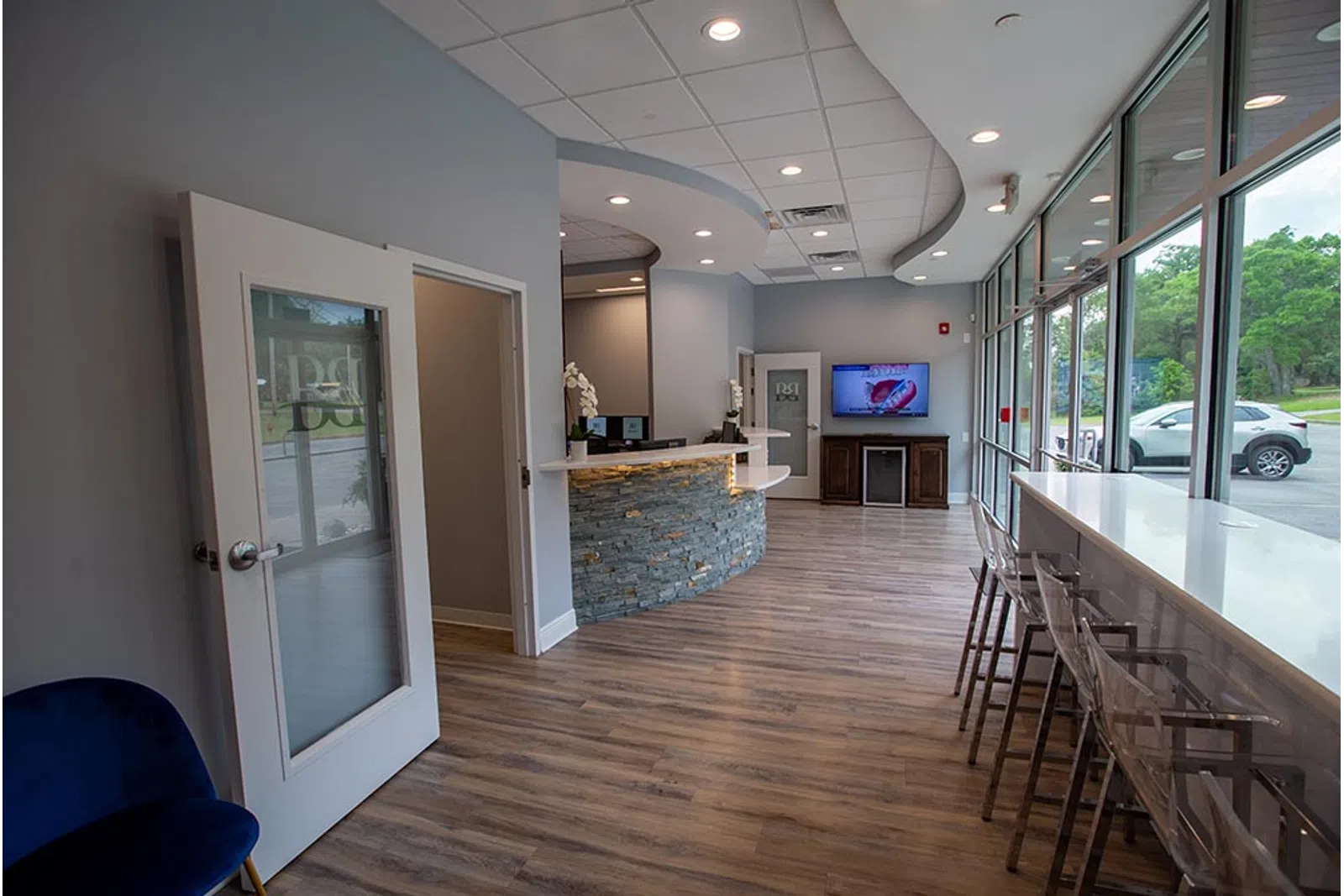 Modern dental office reception area with curved stone desk, wood floor, and large windows with bar stools.