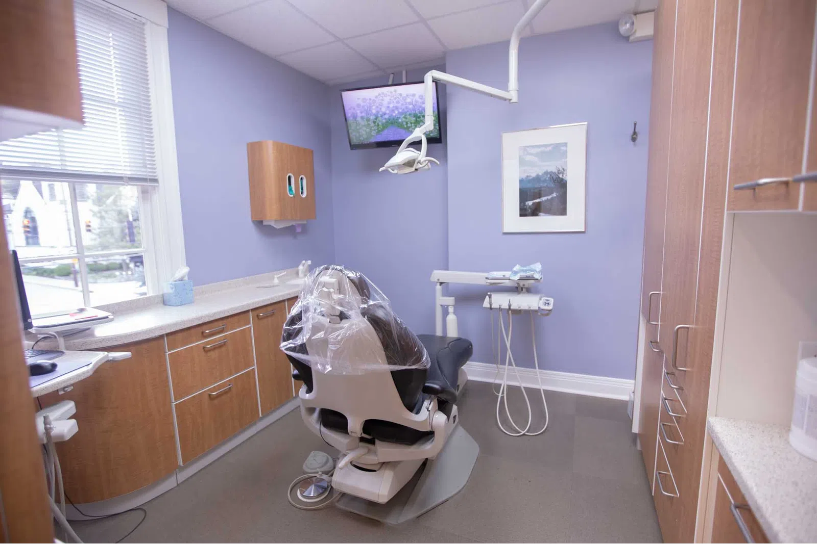 Modern dental office with patient chair, overhead light, cabinets, and monitor on soft purple walls under ceiling lights.