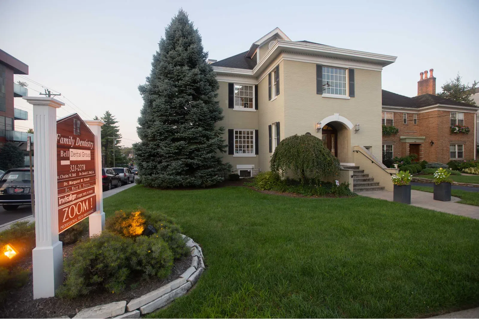 Two-story beige house with black shutters and a dental office sign in a landscaped green lawn.