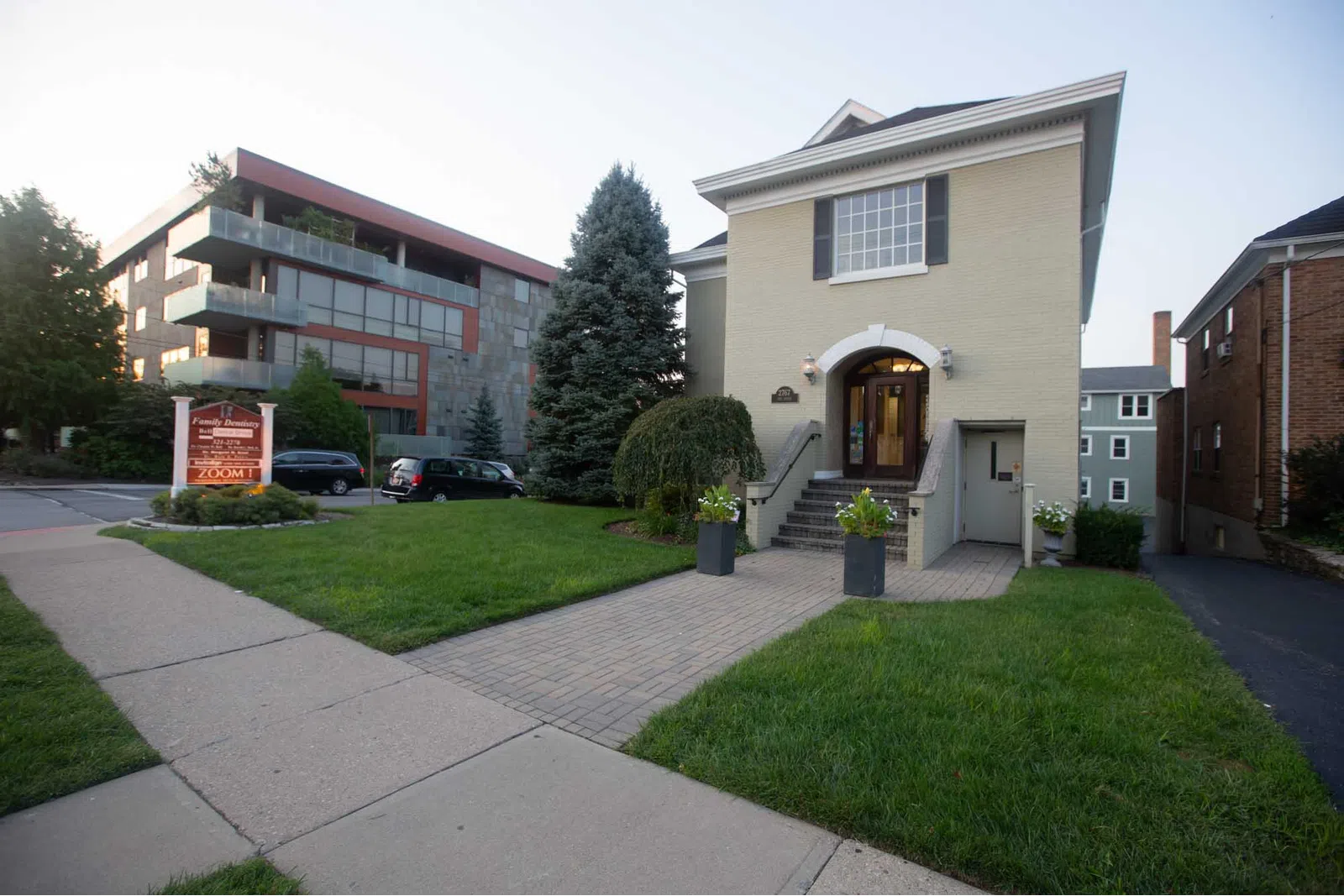 Two-story beige building with stairs, green lawn, sidewalk, trees, and modern apartment in the background.