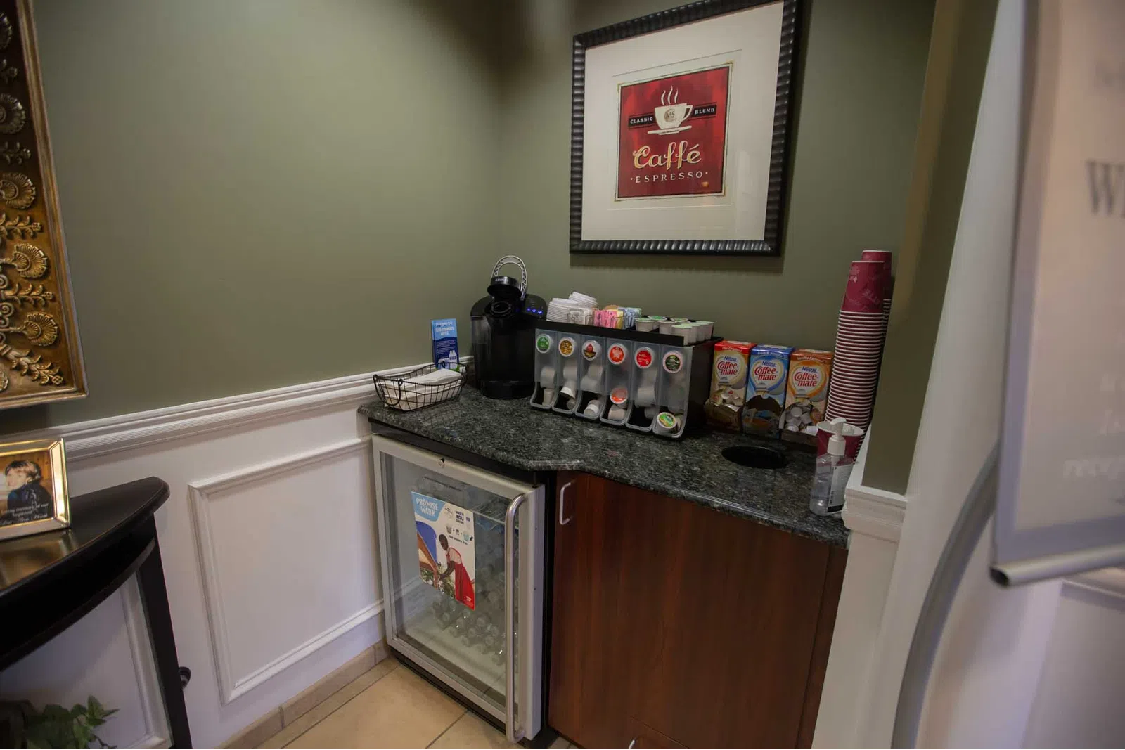 Small coffee station with espresso machine, condiment dispensers, cups, and mini fridge in corner of room.