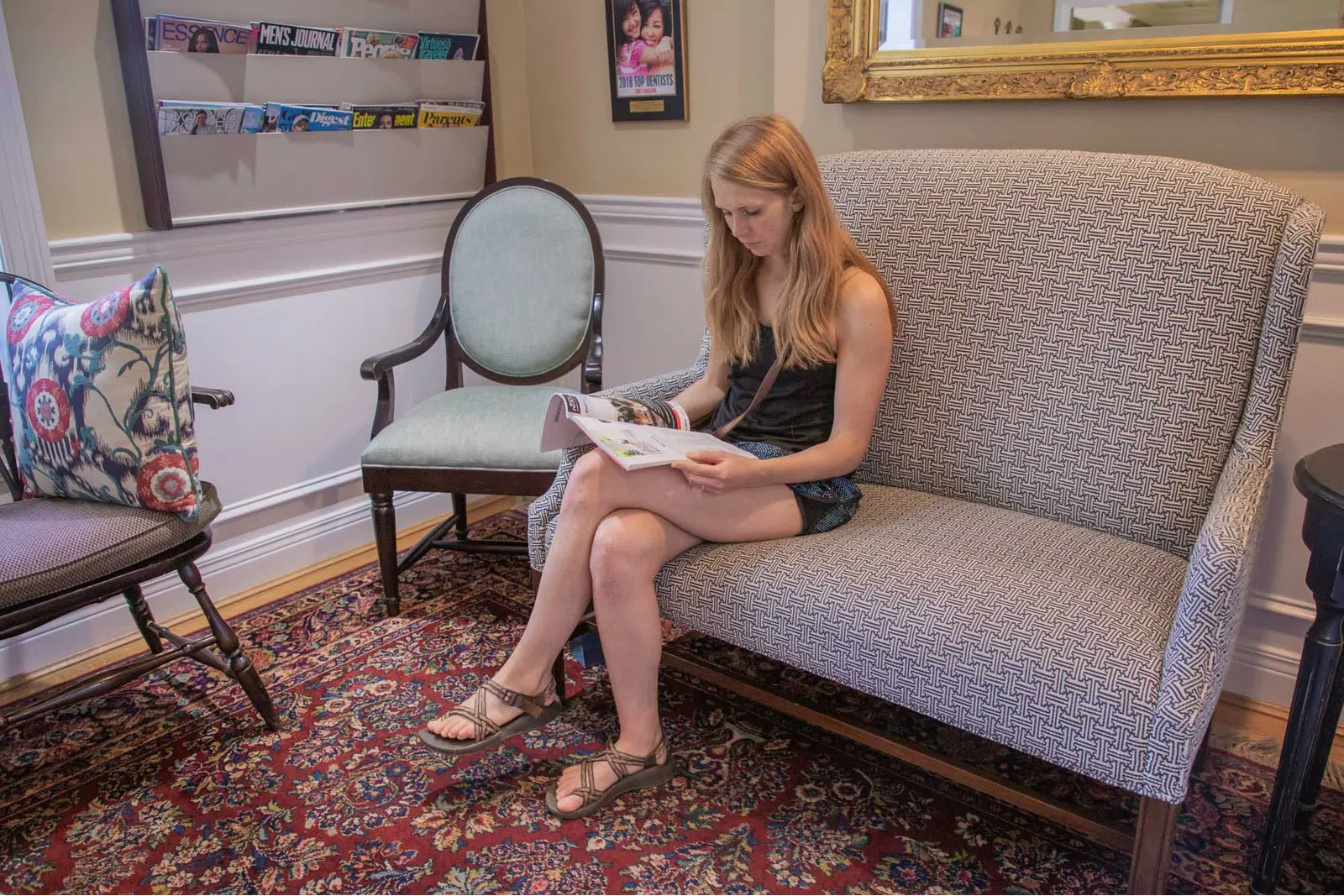 Young woman sitting on patterned sofa reading a magazine in a cozy waiting room with vintage furniture and colorful rug.
