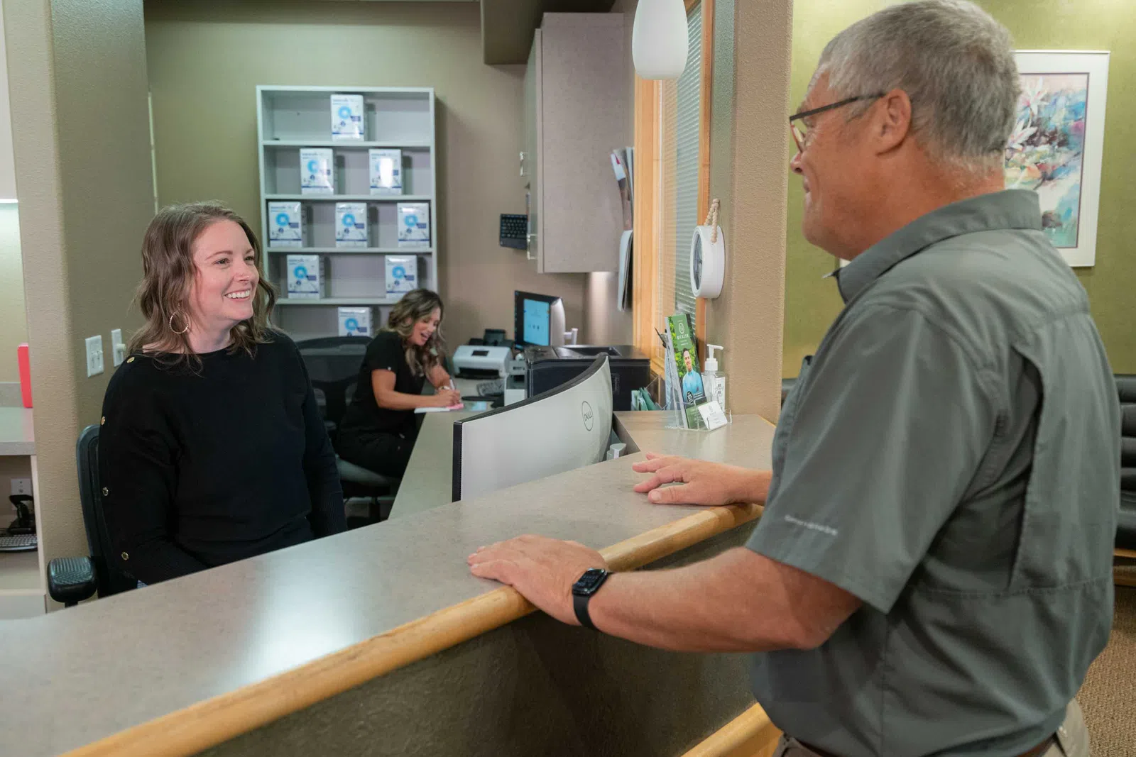 Smiling receptionist assisting an older male client at a medical office front desk with a colleague working in the background
