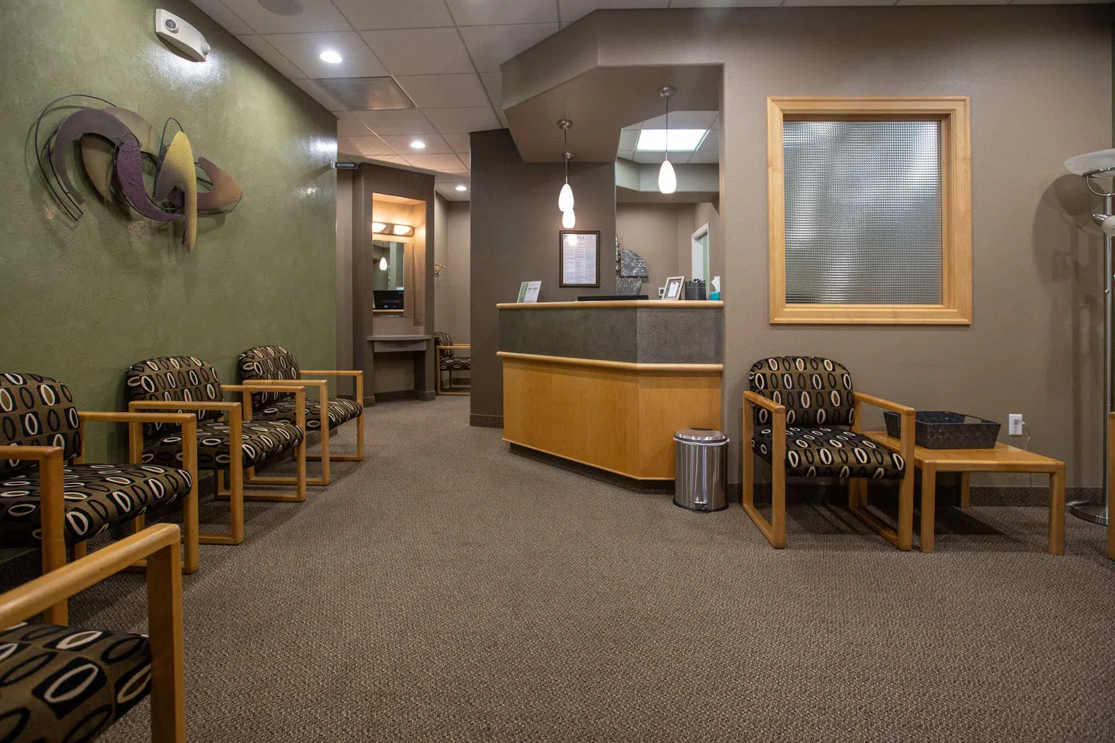 Modern waiting room with patterned chairs, wooden tables, reception desk, and neutral beige walls.
