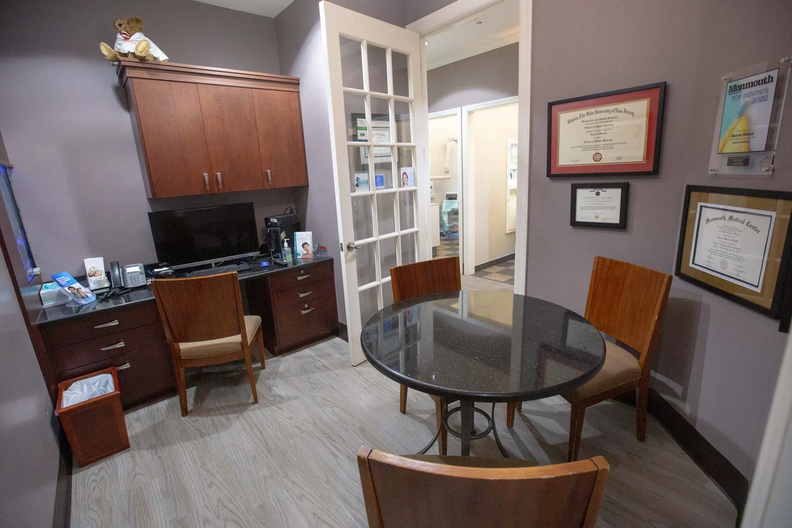 Small professional office with round table, three chairs, computer desk, certificates on gray walls, and wood cabinetry.