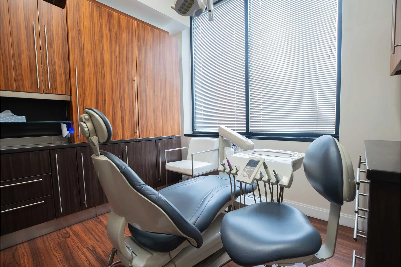 Modern dental clinic room with dental chair, equipment, wooden cabinets, and large window with blinds.