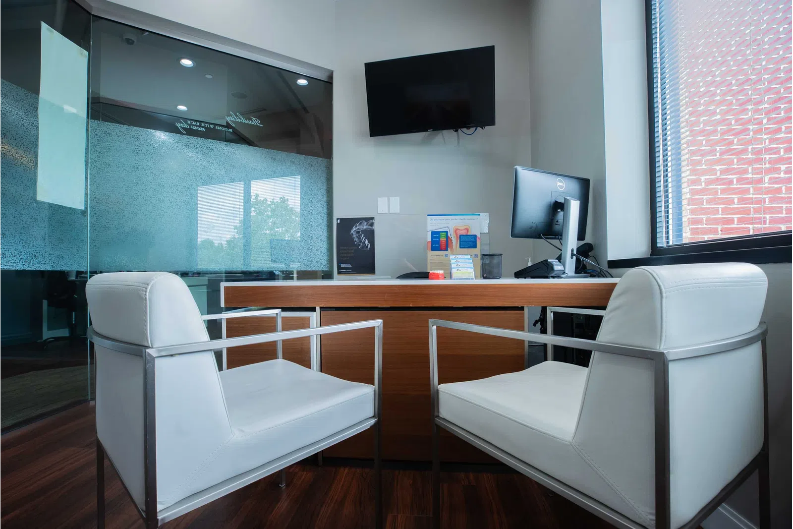 Modern office with two white chairs facing a wooden desk, computer monitor, TV, and large window with blinds.