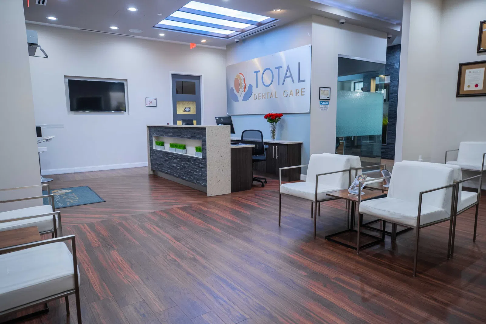 Modern dental office waiting area with white chairs, wood flooring, reception desk, and Total Dental Care sign.