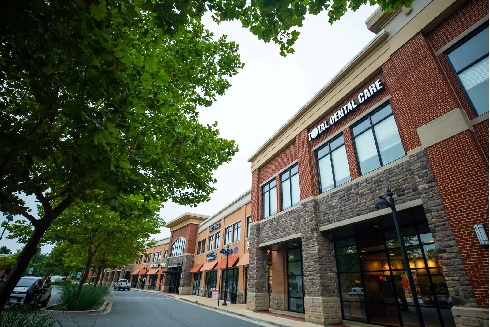 Modern commercial building with Total Dental Care and other storefronts under green trees on a cloudy day.