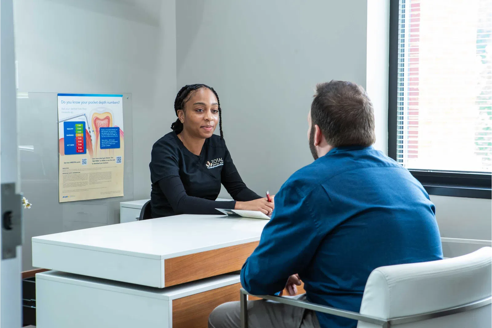 Female healthcare professional consulting a male patient in a modern office with dental health poster visible.