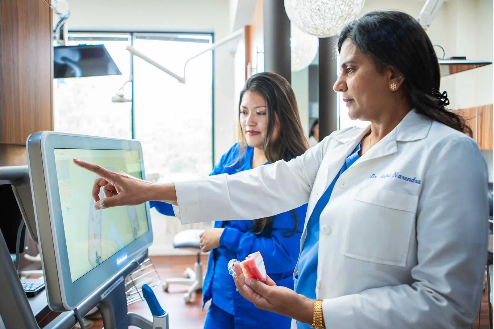 Female dentist in white coat showing dental model and pointing at screen with assistant in clinic.
