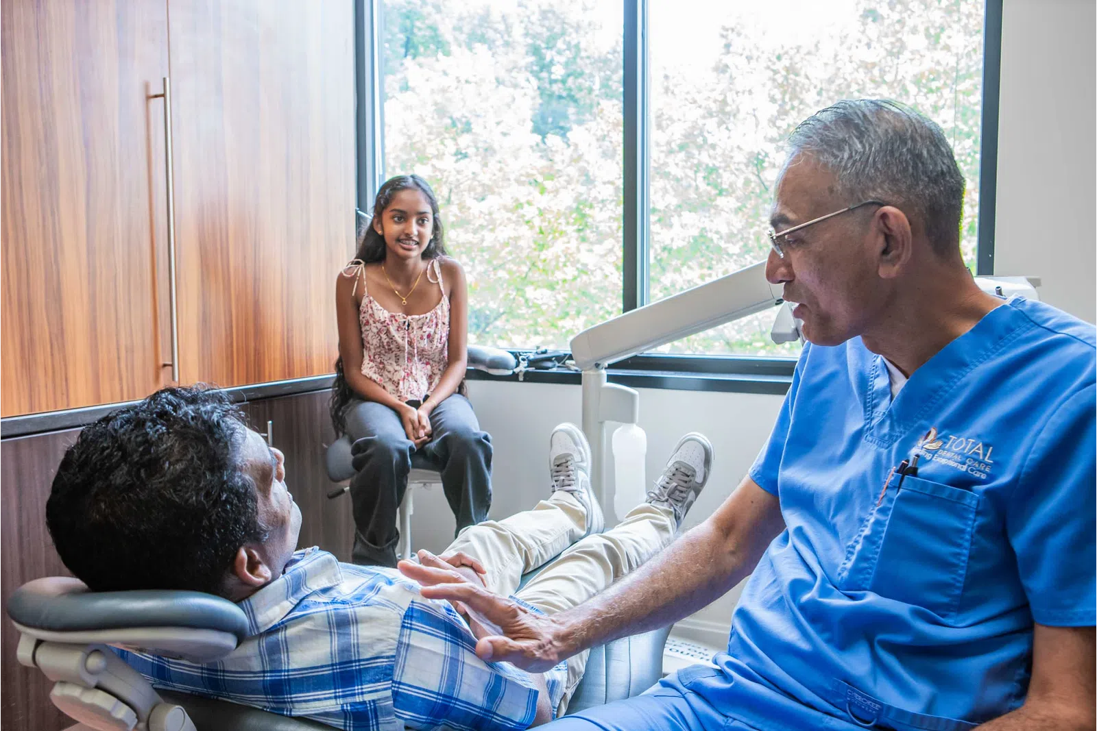 Dentist in blue scrubs talking to male patient in dental chair while young girl sits nearby by window.