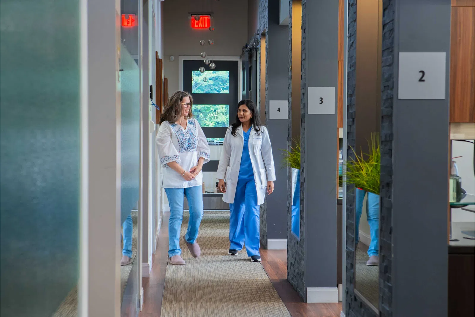 Doctor and patient walking and talking together in a modern medical clinic hallway with numbered rooms.