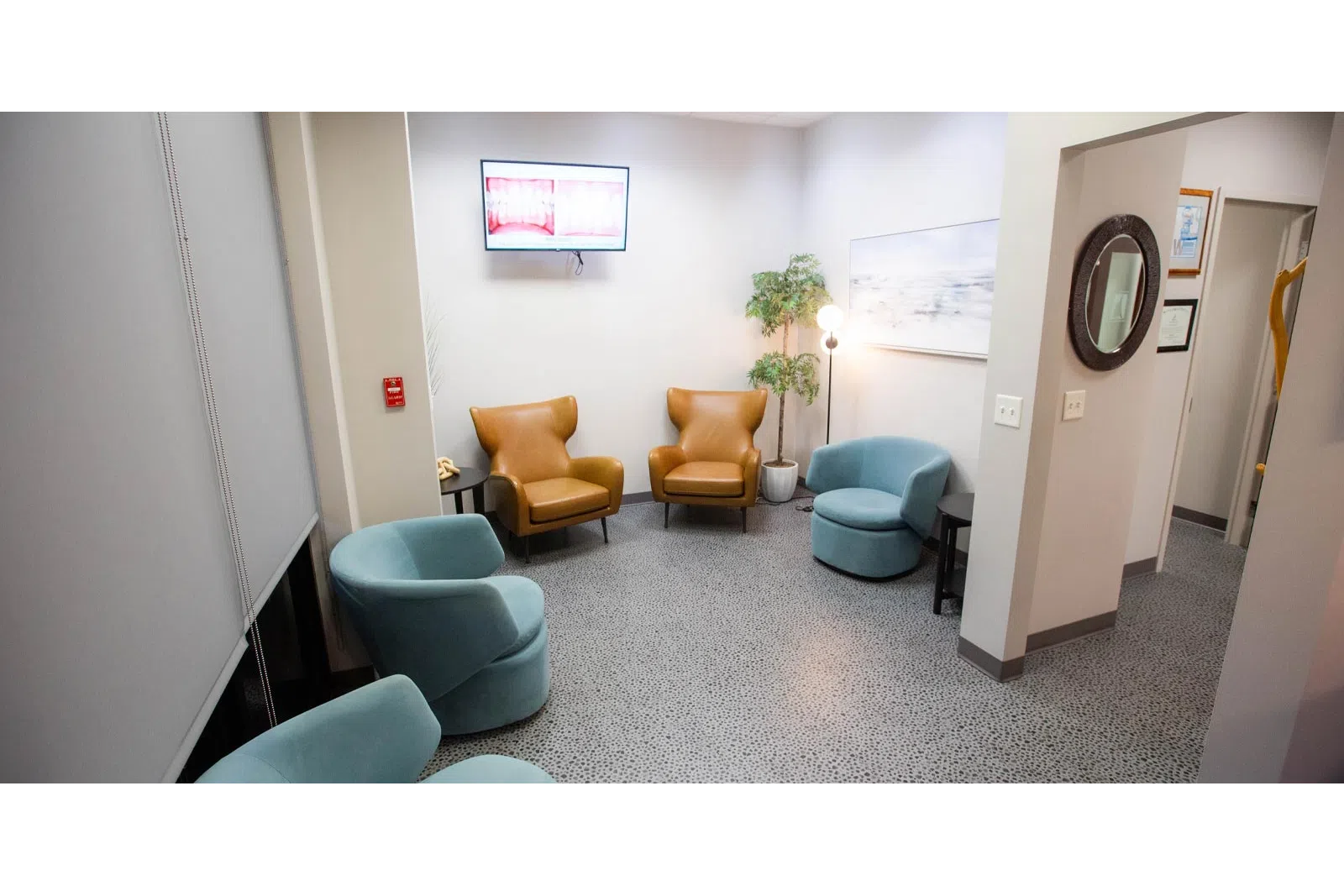 Modern waiting room with brown and blue chairs, a plant, floor lamp, and a wall-mounted TV screen.