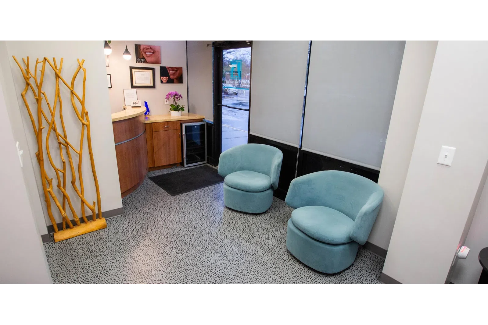 Modern waiting area with two blue chairs, wooden reception desk, decorative wooden sculpture, and window blinds.