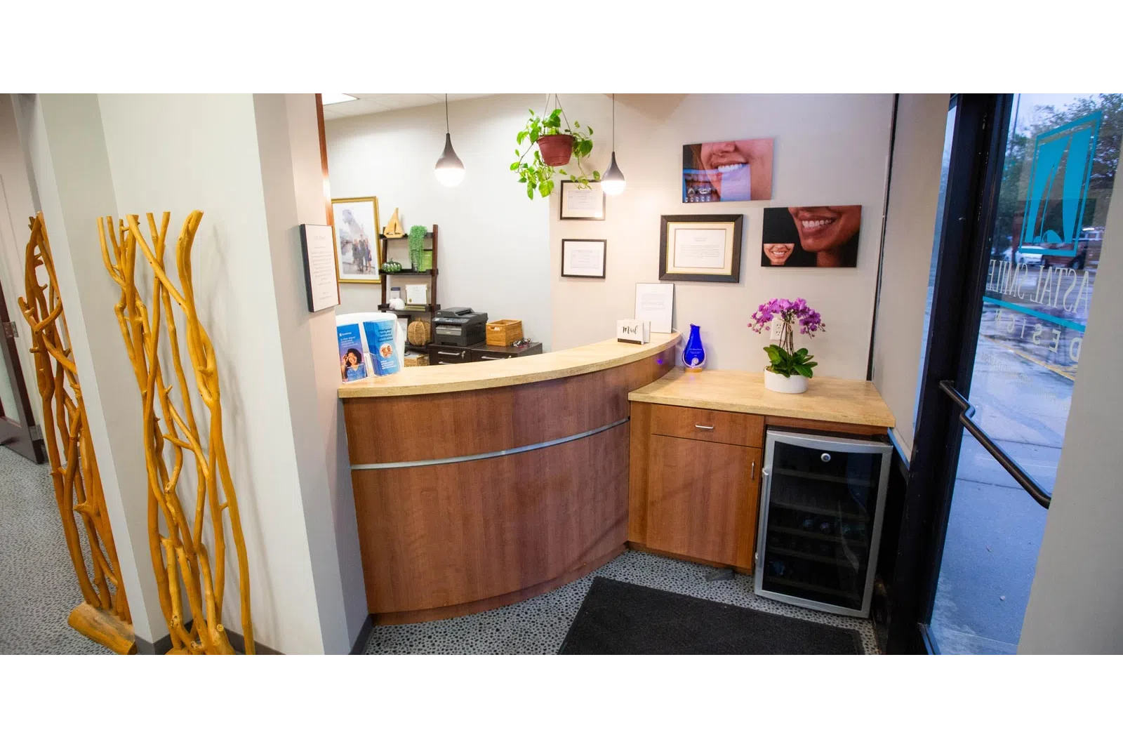 Reception area with curved wooden desk, plants, framed certificates, and a small refrigerator near glass entrance door.
