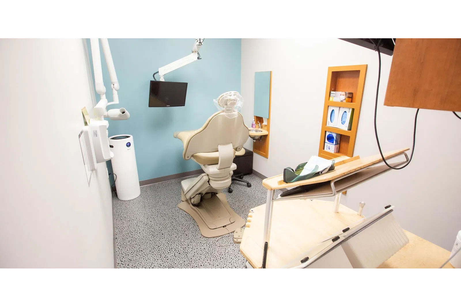 Modern dental clinic room with beige dental chair, overhead light, TV screen, and dental equipment.