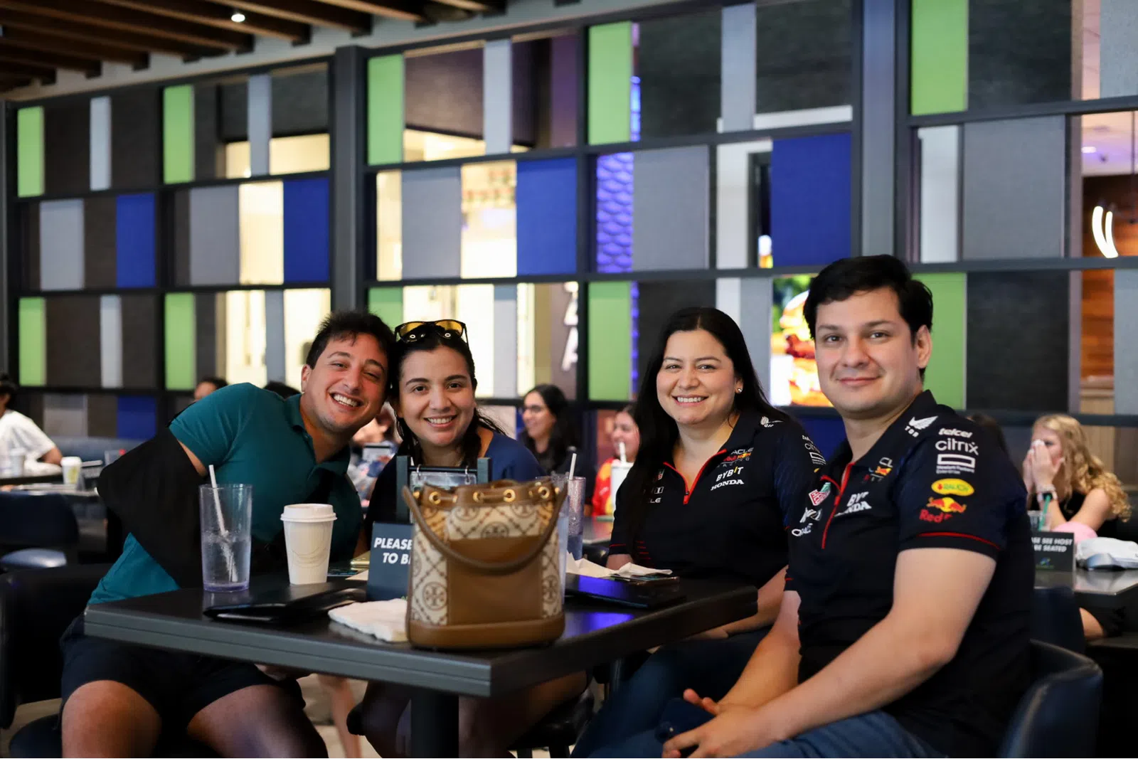 Four friends smiling together at a bustling café, enjoying drinks and each other's company.