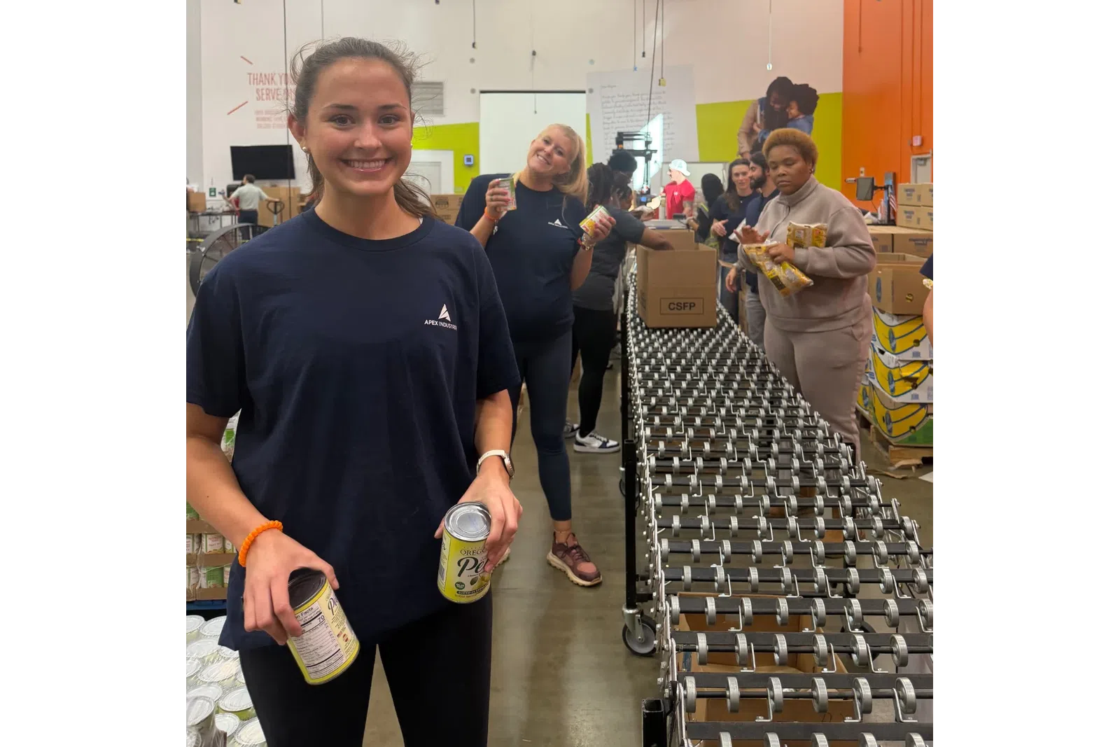 Volunteers packing canned food items on a conveyor belt in a busy warehouse setting