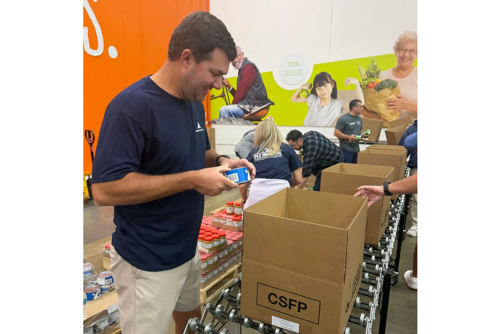 Volunteers packing food items into CSFP boxes at a warehouse with a mural in the background.