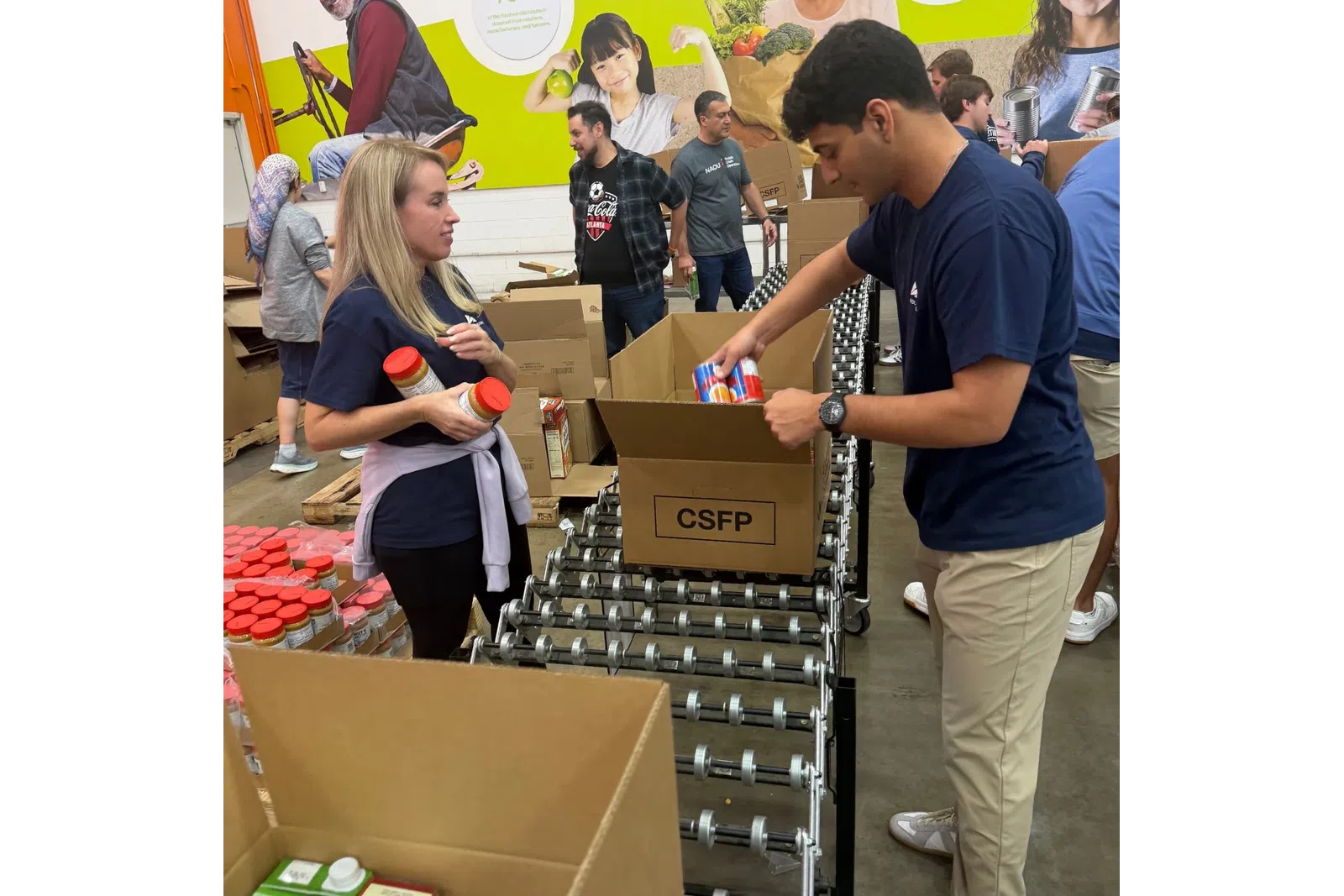 Volunteers packing canned goods and jars into CSFP boxes on a conveyor in a food bank with mural background.