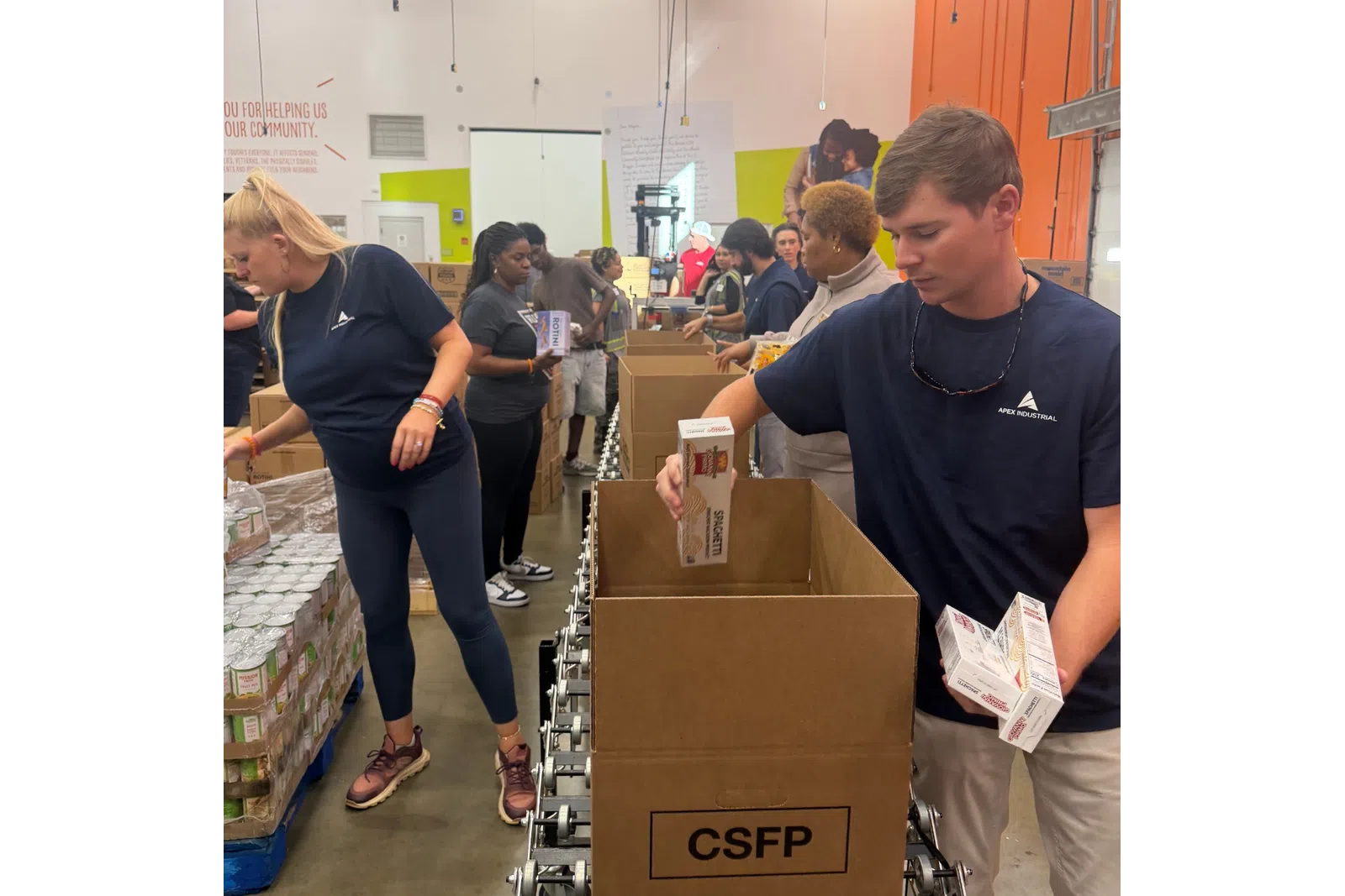 Volunteers packing food boxes with pasta and canned goods at a community food bank warehouse.