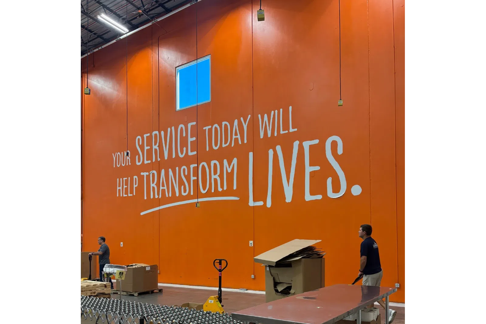 Workers handle pallets and cardboard in a warehouse with an orange wall and overlaid text saying your service today will help transform lives.