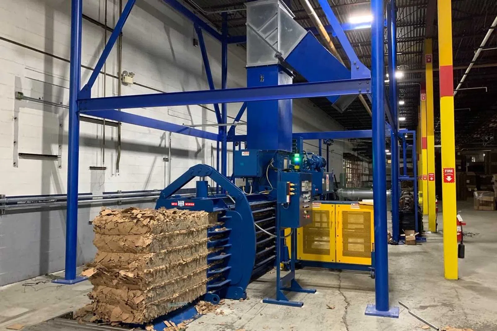 Industrial cardboard baler compressing cardboard into large stacks inside a warehouse environment.