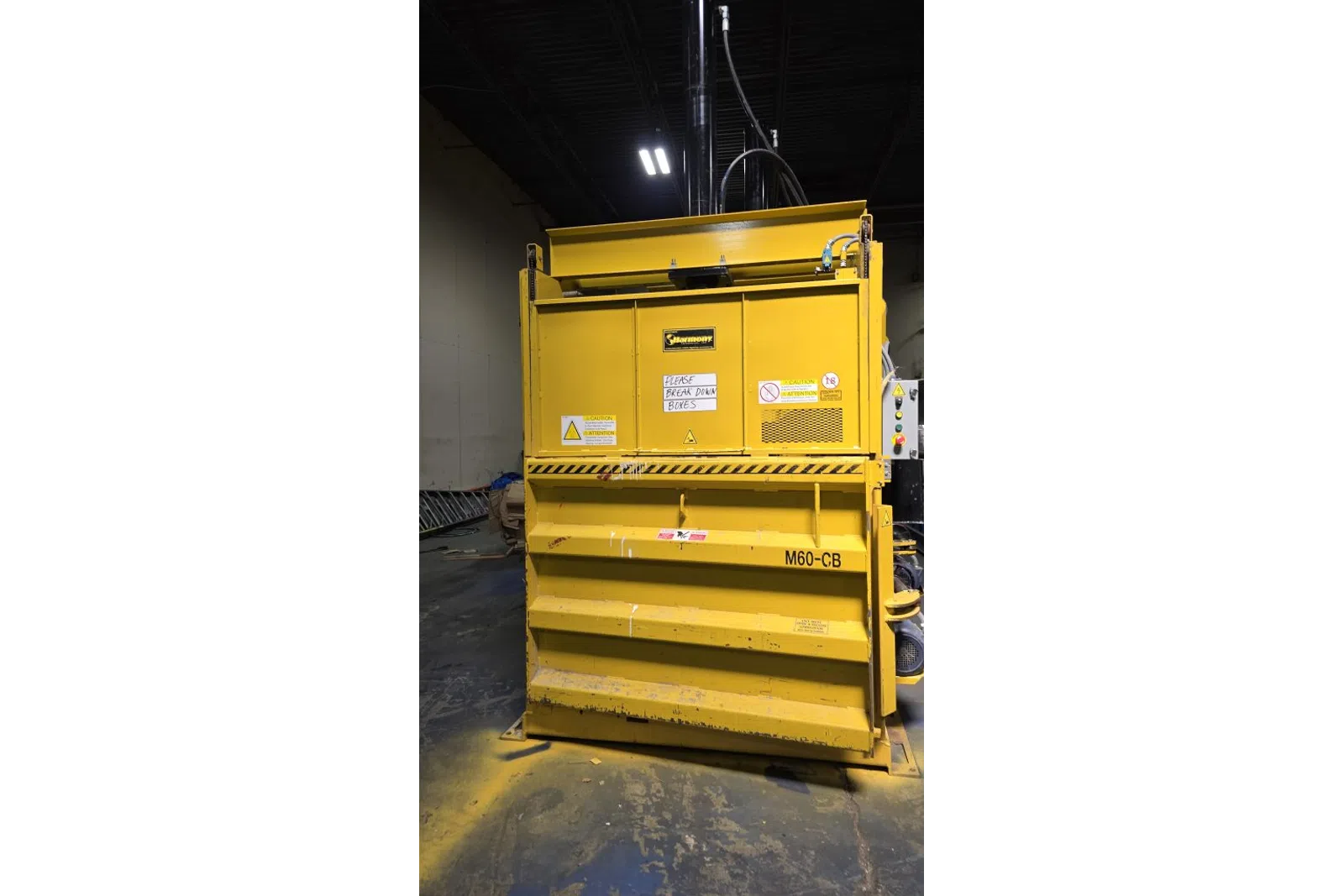Large yellow industrial baler machine labeled M60-CB in a warehouse with caution signs and instructions.