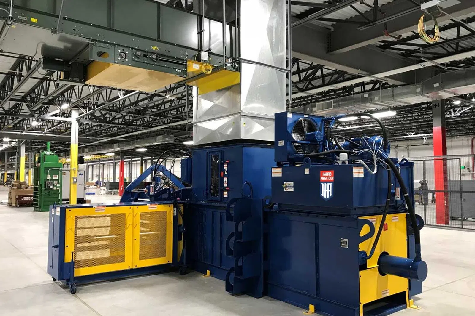 Large blue industrial machine with yellow safety doors inside a spacious factory with metal beams and ventilation ducts.
