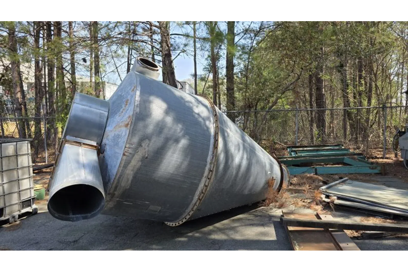 Large metal industrial silo lying on its side in an outdoor storage area surrounded by trees and equipment.