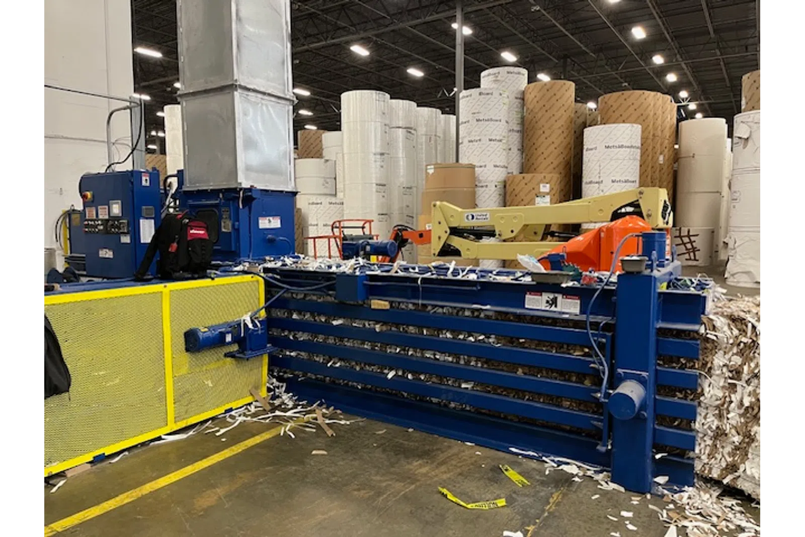 Industrial paper baler and large rolls of paper stacked in a warehouse with equipment and safety barriers.