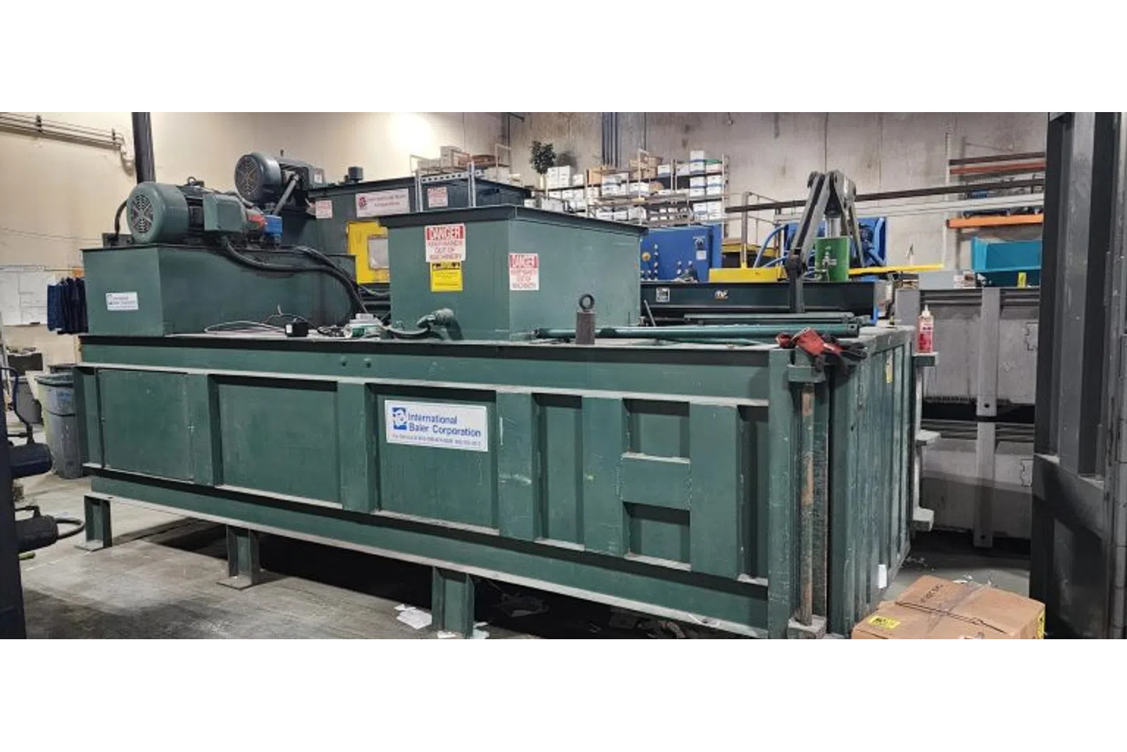 Large green industrial baler machine inside a warehouse with warning labels and company signage.