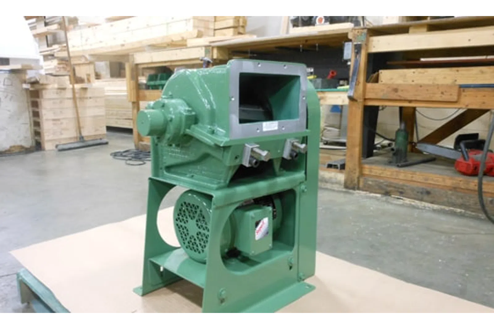 Green industrial machine with motor, mounted on a base in a workshop with wood and tools in the background