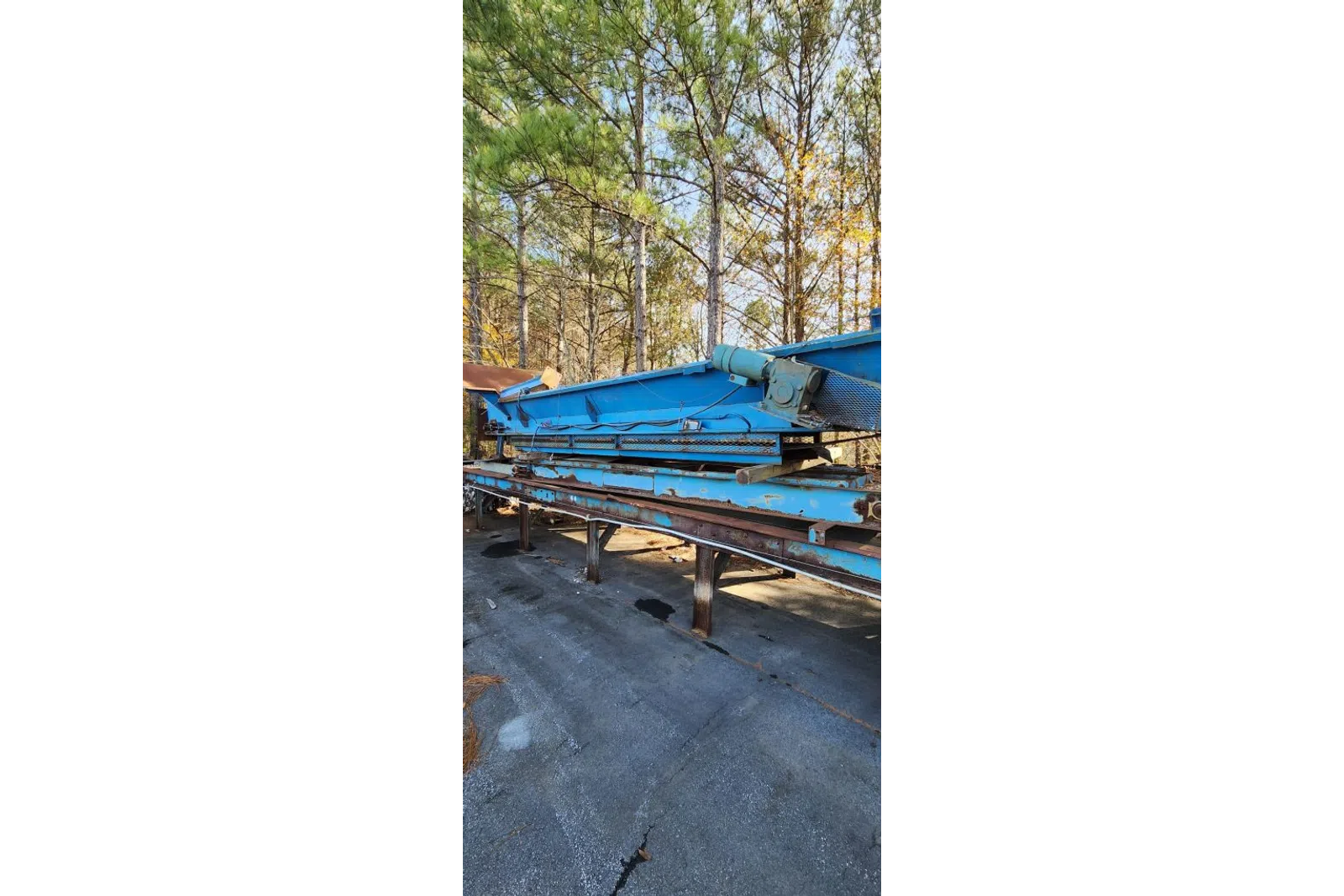 Large blue industrial conveyor belt machine outdoors near trees on cracked asphalt surface.
