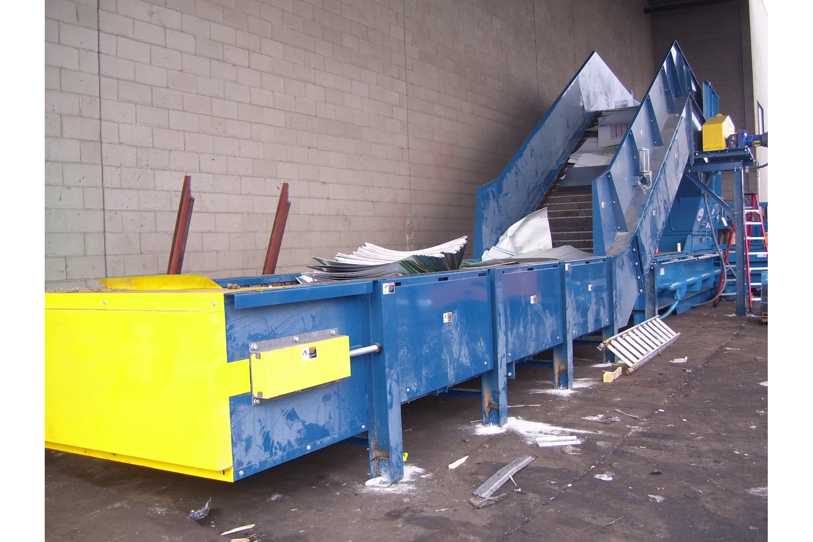 Industrial blue and yellow conveyor belt system inside warehouse with scattered debris on floor.