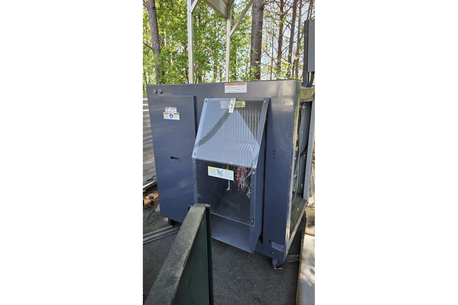 Large industrial gray machine with a transparent window panel outdoors under a canopy near trees.