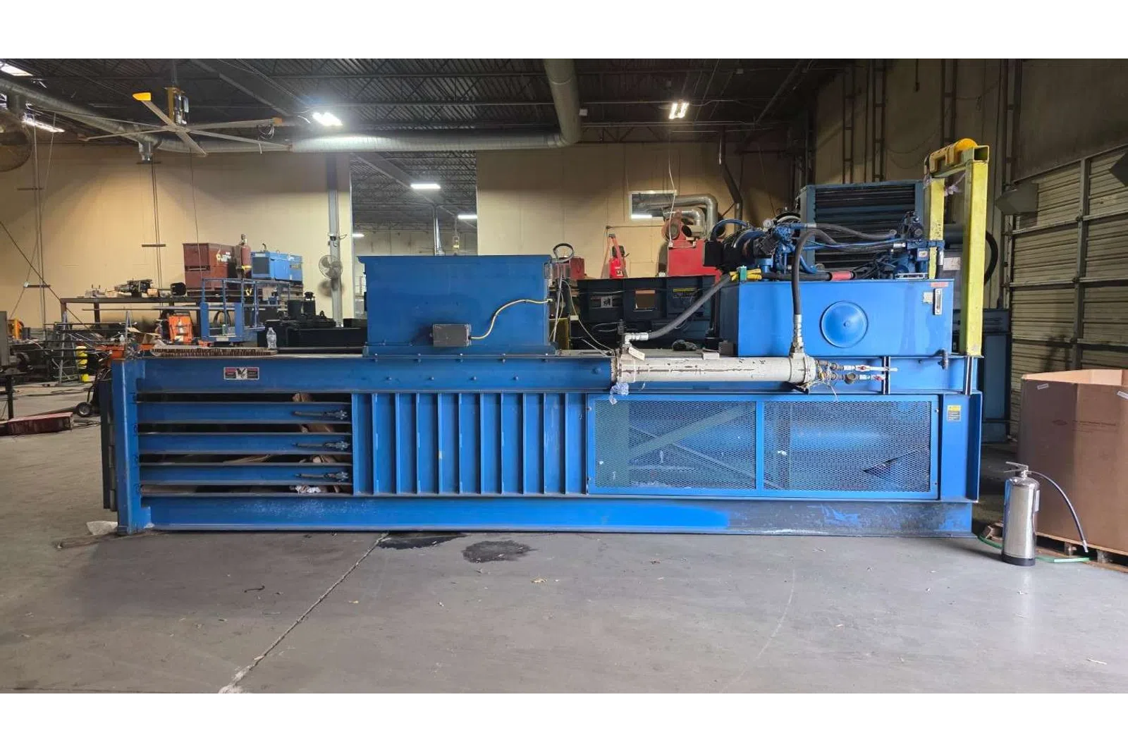 Large blue industrial baler machine inside a warehouse with concrete floor and metal shelves nearby.