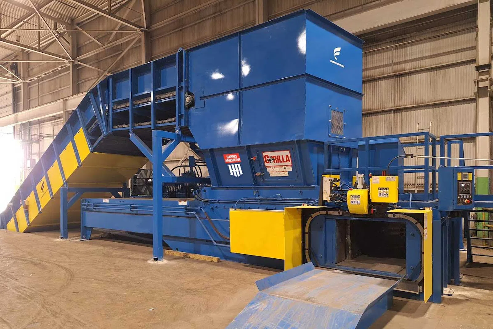 Large blue and yellow industrial baling machine in a factory warehouse setting with conveyor belt and control panels.