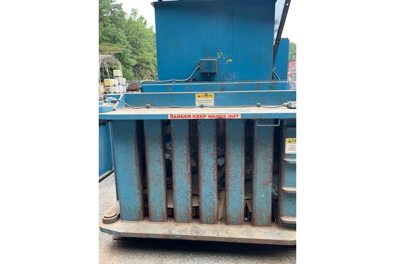Rusty blue industrial trash compactor with caution signs and visible compressed waste inside.