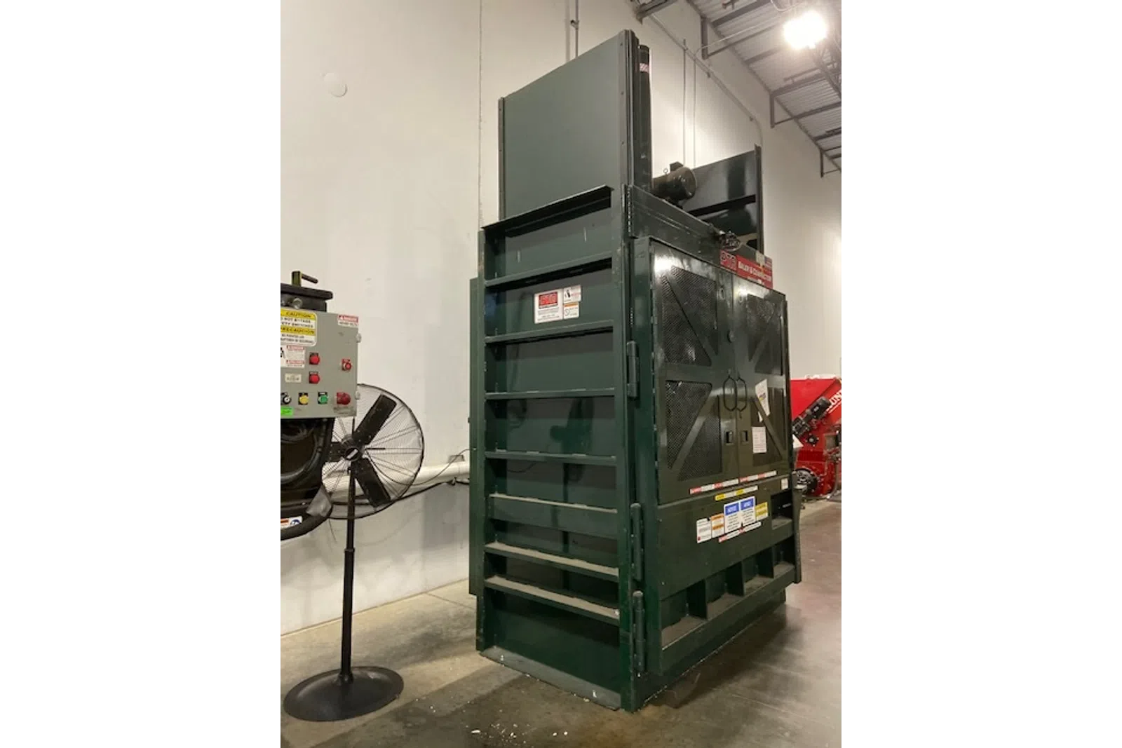 Large green industrial baler machine inside warehouse with control panel and standing fan nearby