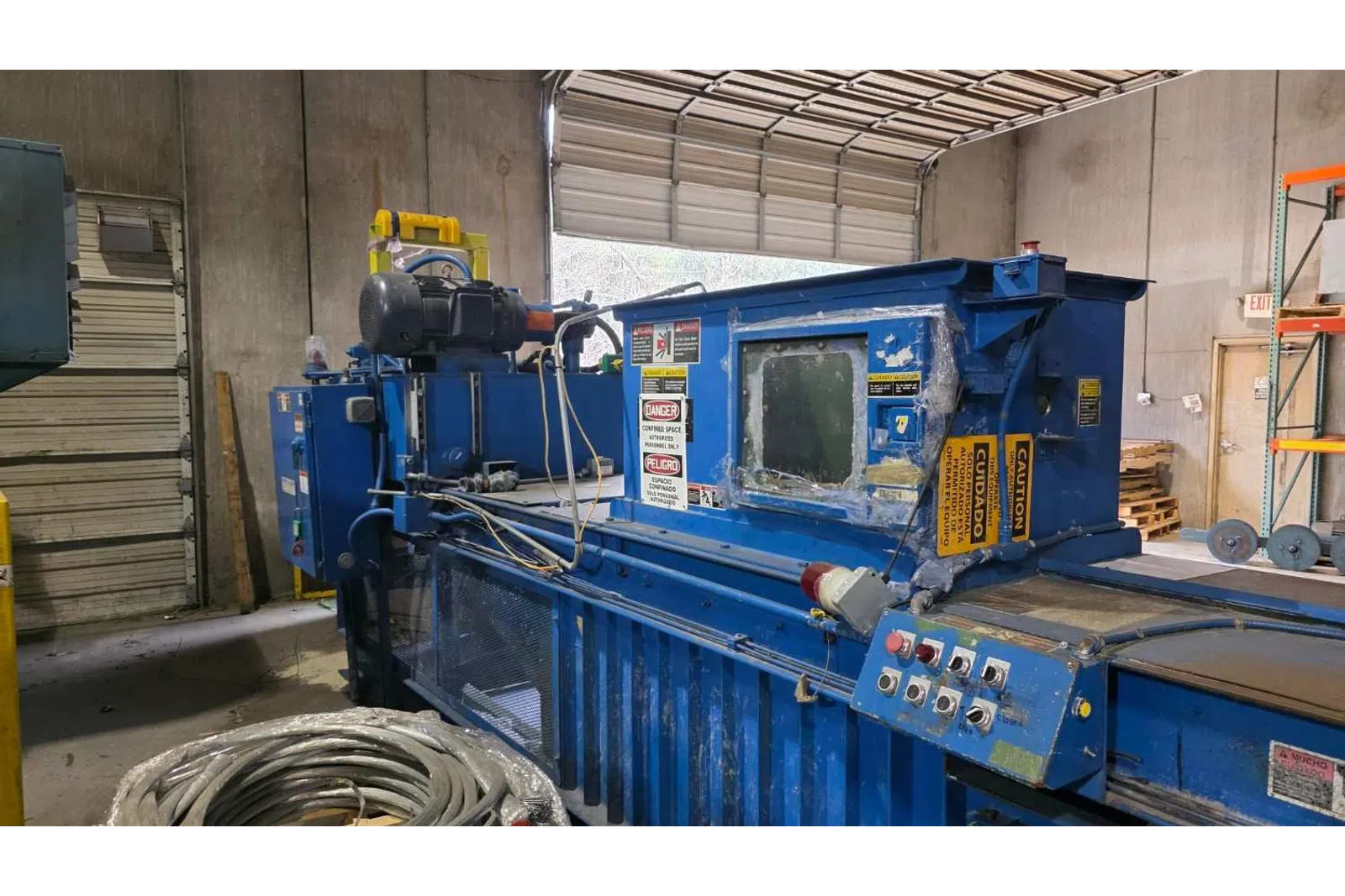 Blue industrial machine with control panel inside a warehouse near coiled cables and open garage door.