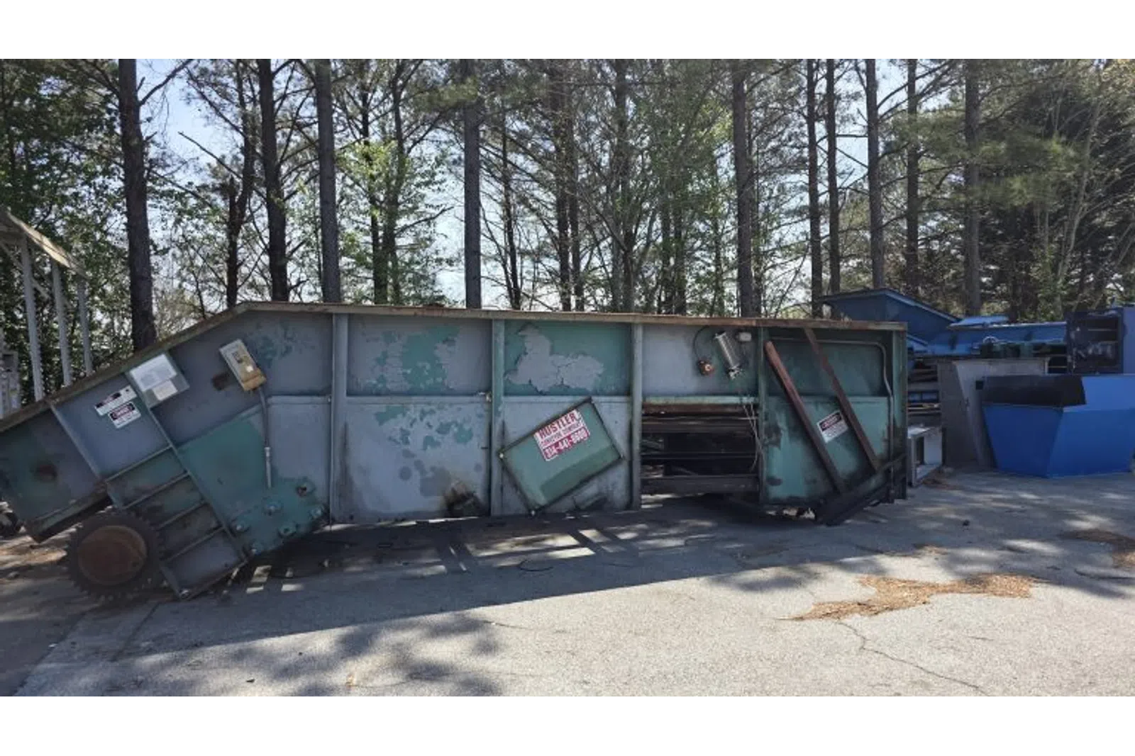 Old industrial metal machinery with rust and peeling paint outdoors near trees and blue equipment.