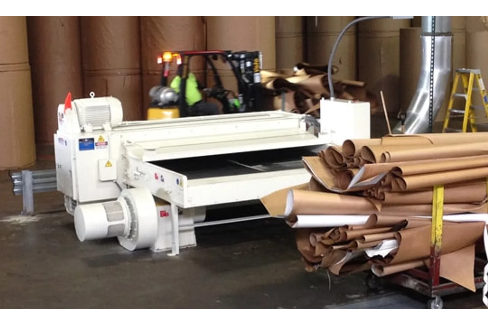 Industrial paper manufacturing equipment with large rolls of brown paper and forklift in warehouse.