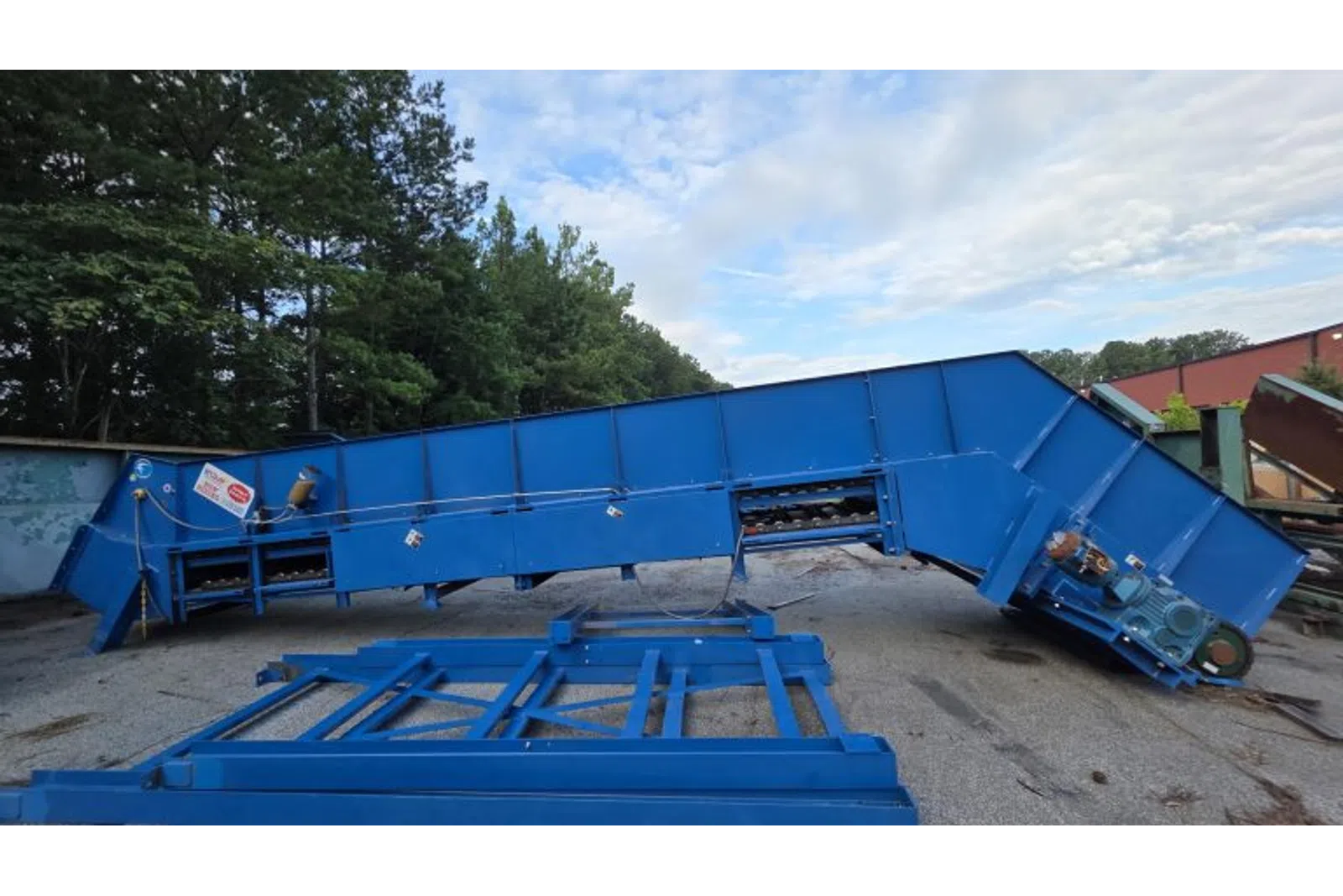 Large blue industrial conveyor belt system outdoors on gravel near trees and buildings under partly cloudy sky.