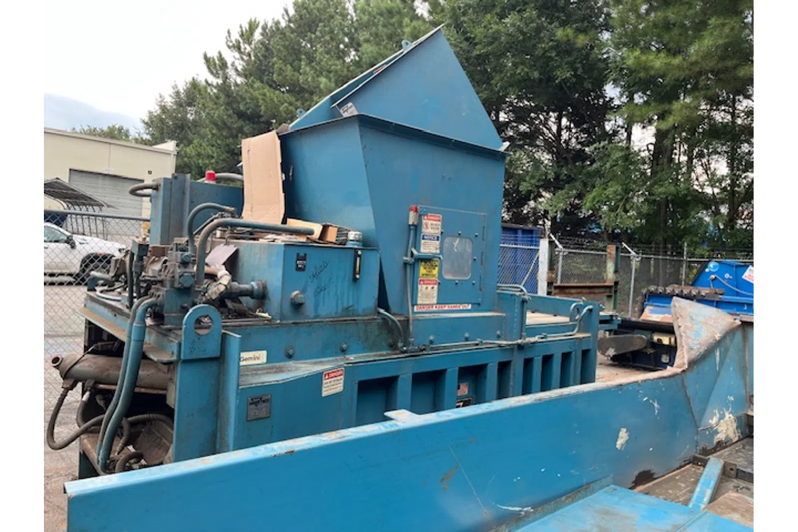 Large blue industrial machine with hydraulic components and chute in outdoor fenced area near trees and vehicles.
