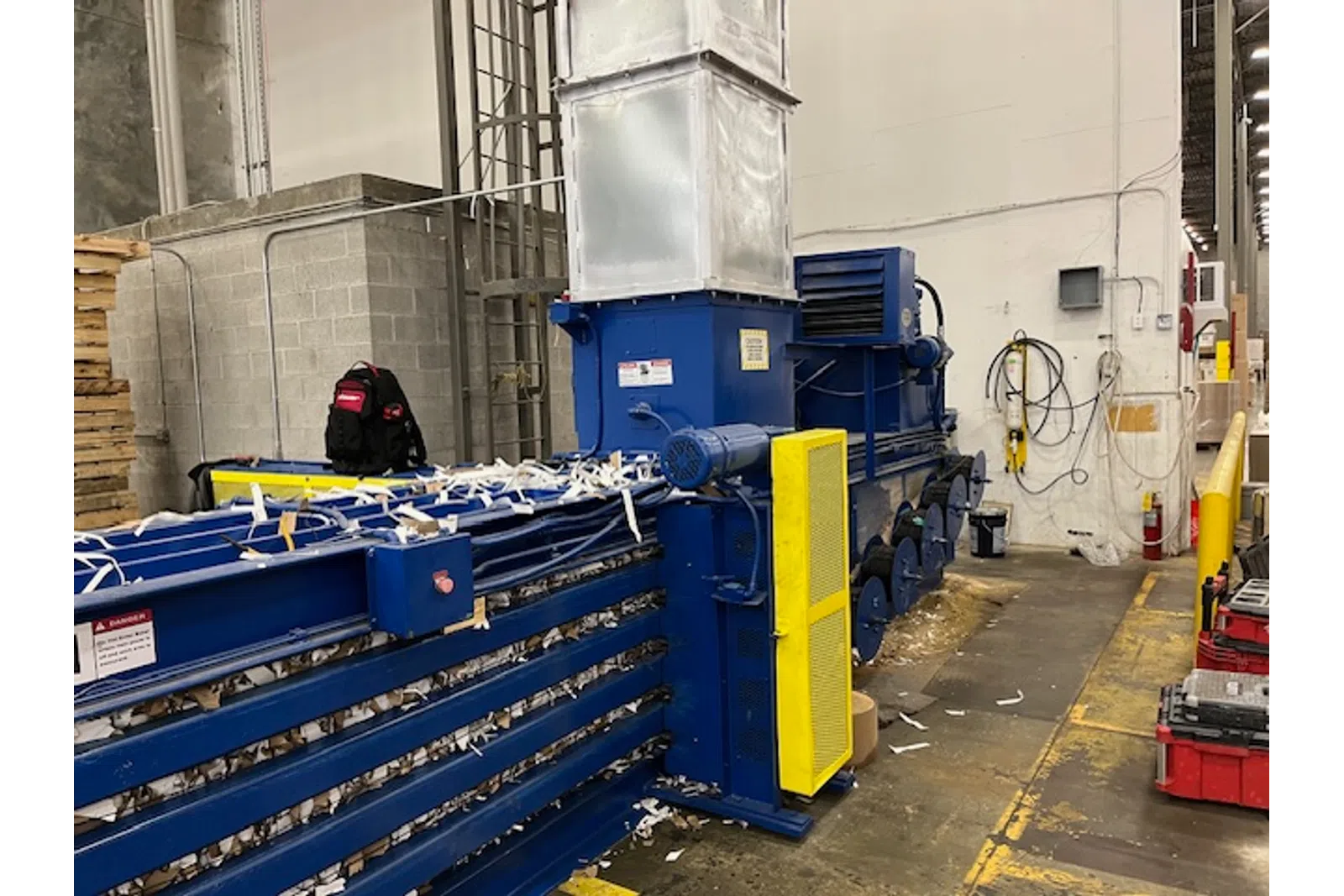 Large blue industrial baler machine compressing shredded paper inside a warehouse with concrete walls and yellow safety barriers.