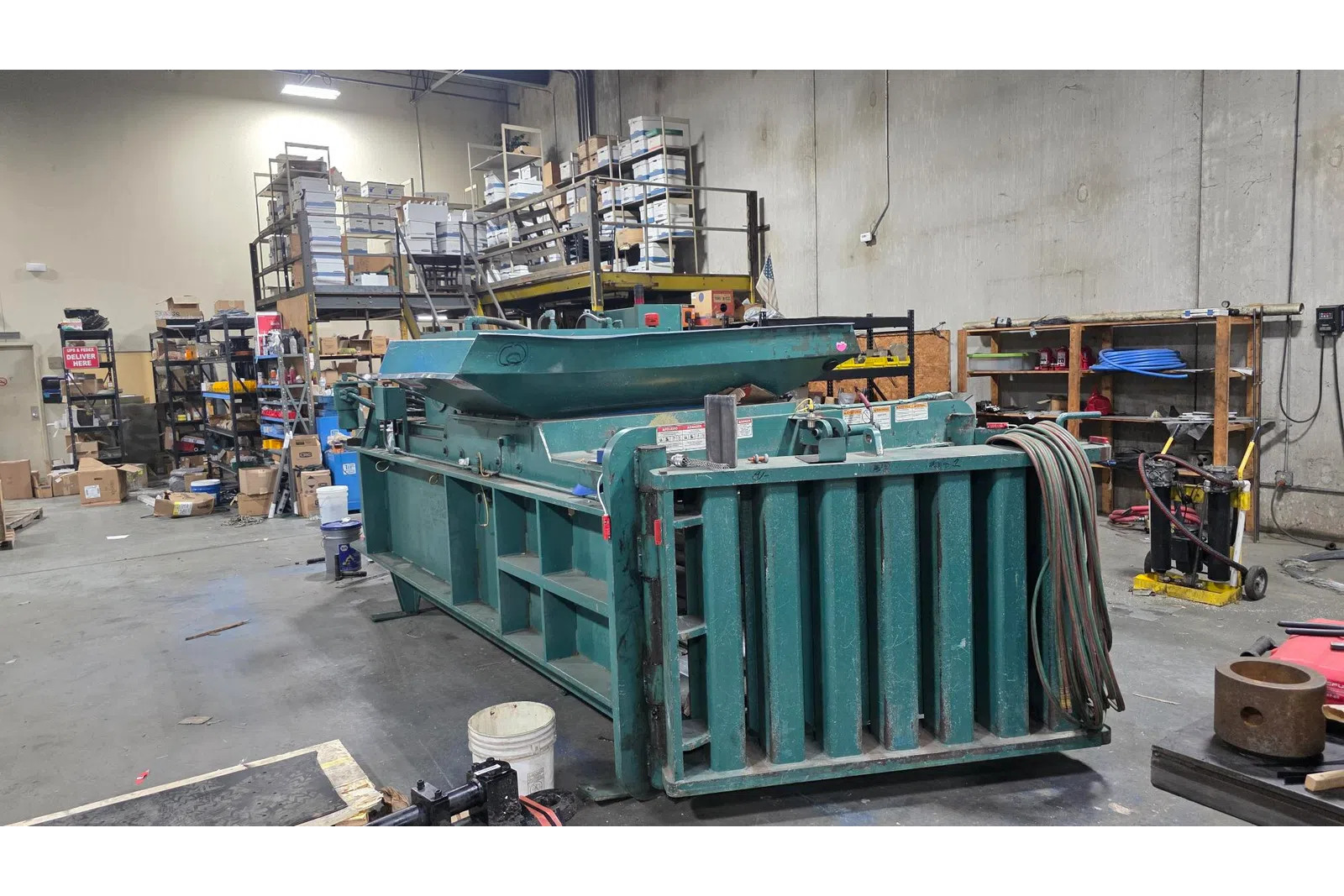 Large industrial green metal baler machine inside a cluttered warehouse with shelves and boxes.