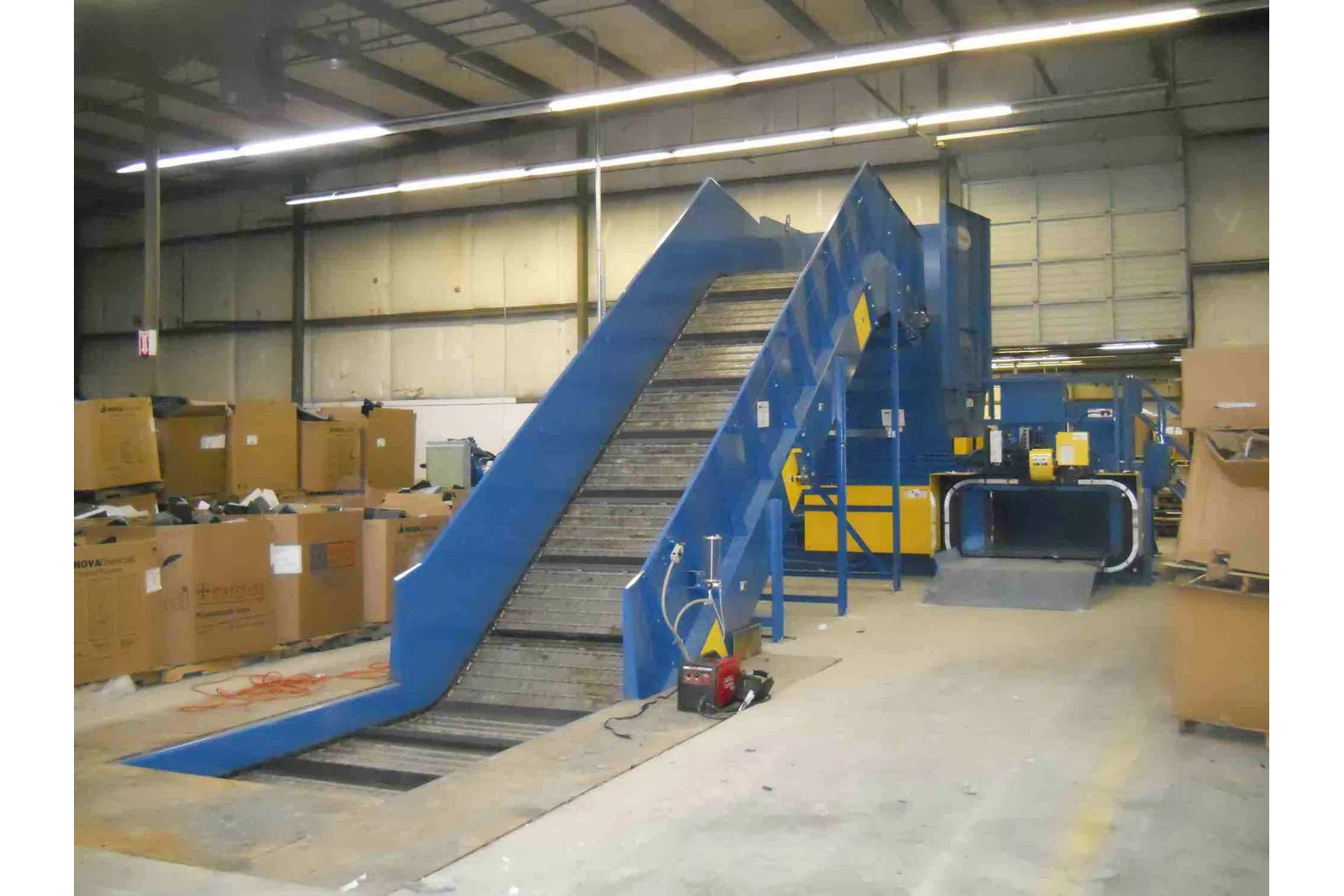 Blue industrial conveyor belt system in warehouse surrounded by cardboard boxes and equipment