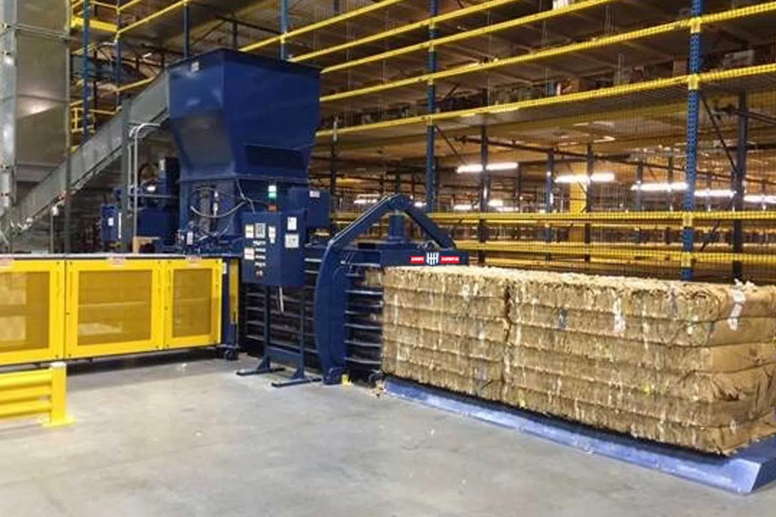 Automated industrial baler compressing cardboard bales in a warehouse with yellow safety barriers and metal racks.
